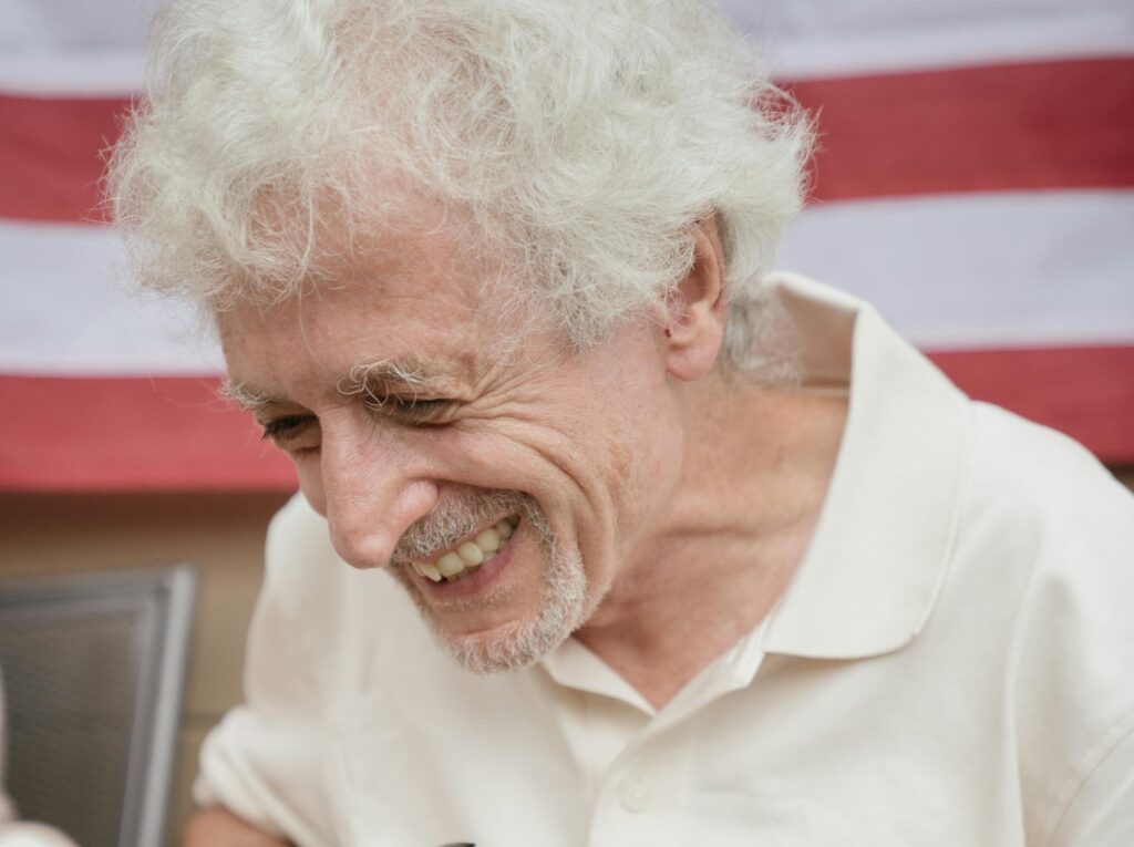 Senior man joyfully opening a wine bottle outdoors with an American flag in the background.